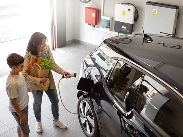 A woman plugging in an electric car for charging in a garage, with a child standing nearby. Solar inverters and a battery system are visible on the wall.